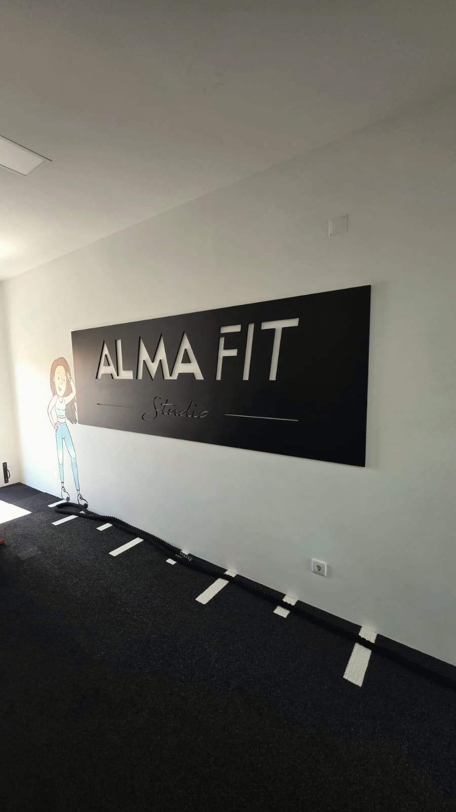 Corredor de treino do estúdio com a placa AlmaFit Studio na parede e o chão marcado.